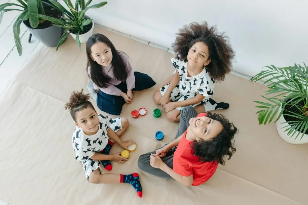 Four diverse children sit happily on the floor painting together indoors.