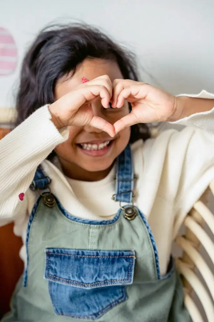 Anonymous cheerful Hispanic preschool girl with sticker on forehead making heart shaped hand gesture while sitting near wall with egg decoration during Easter holiday